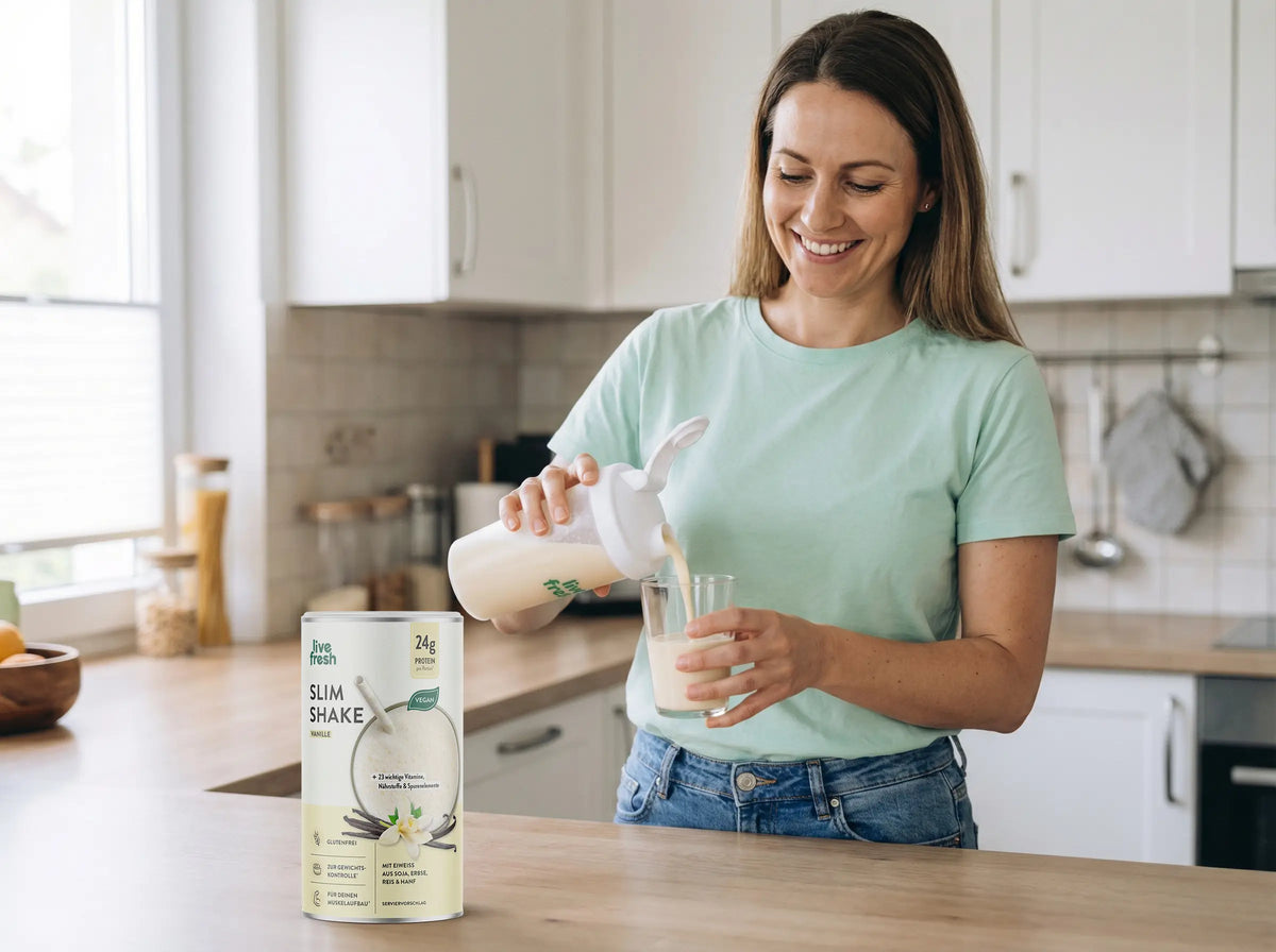 Frau bereitet in einer hellen Küche einen LiveFresh Slim Stoffwechselshake zu und gießt den Shake aus einem Shaker in ein Glas. Daneben steht die Dose Slim Shake Vanille mit 24 g Protein und veganer Rezeptur.