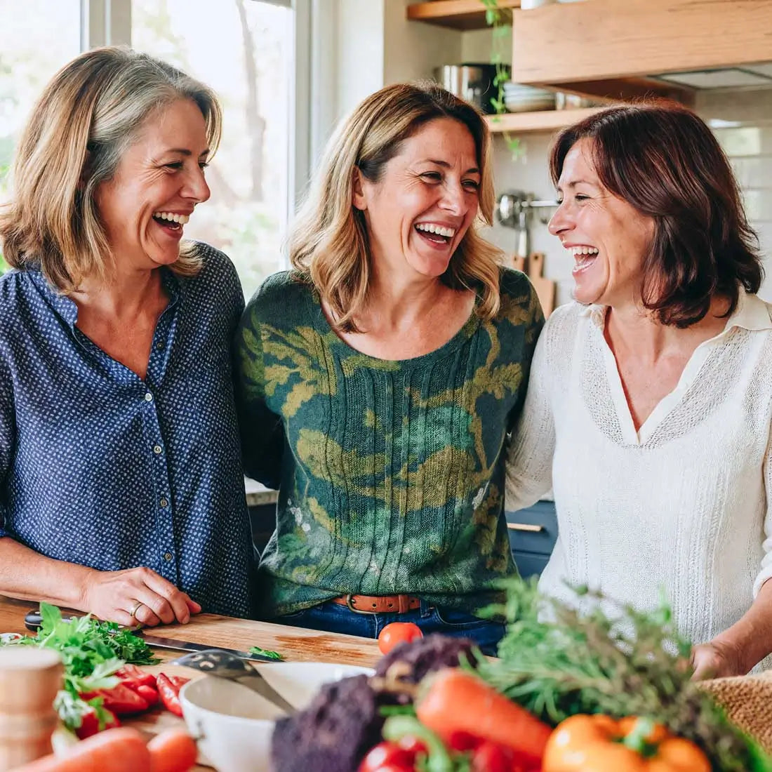 Drei Frauen stehen in der Küche und haben Spaß am Kochen. Im Vordergrund sind frische Zuatten wie Tomate, Salat, Karotte und Kräuter zu sehen